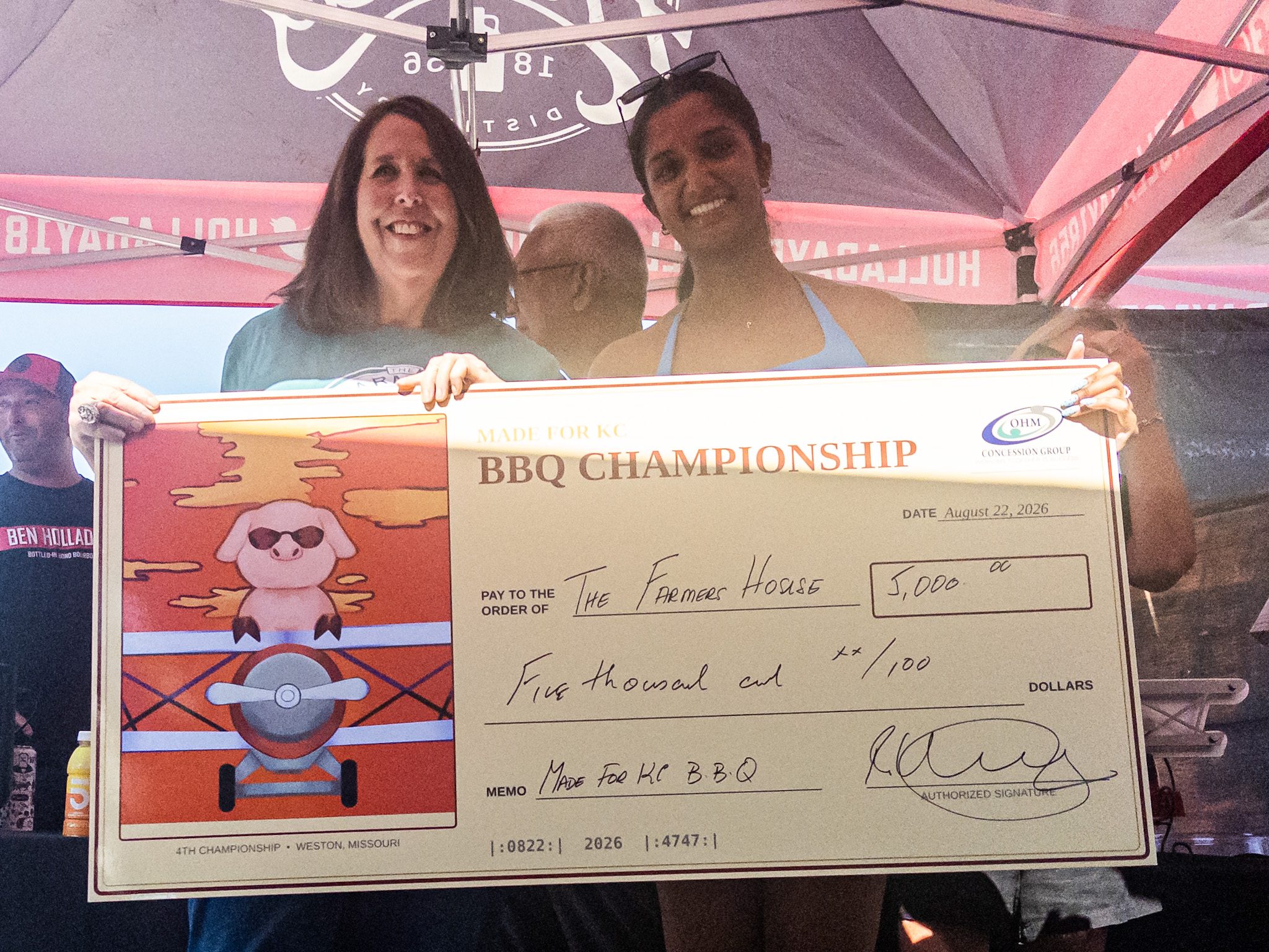 Photo by MEGAN JANTOS
Dana Chatlin, left, director of development and marketing at The Farmer’s House, and a OHM Concession Group representative, hold a ceremonial $5,000 donation check during the Made for KC BBQ Championship awards ceremony Saturday, Aug. 22, outside Weston City Hall in Weston